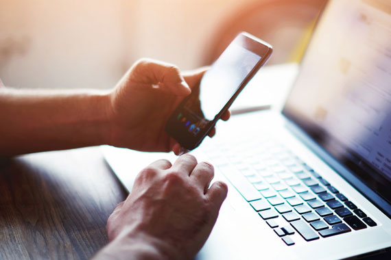 Ansicht von zwei Händen einer Person auf einem Tisch. Die eine Hand hält ein Handy, die andere bewegt den Cursor auf einem Laptop.