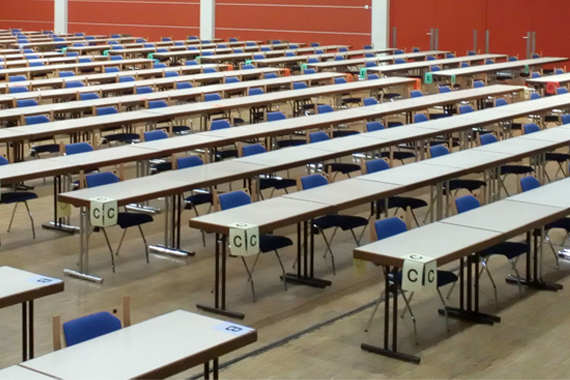 Blick eine große Halle mit Tischen für die Testabnahme. An jedem Tisch steht eine Stuhl. Auf den Tischen kleben Buchstaben, welche die Sektoren im Testraum markieren.